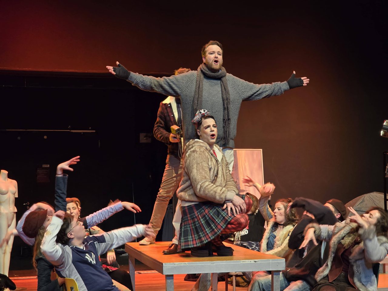 Jelle Gulmans staand op een tafel, zingend te midden van de cast